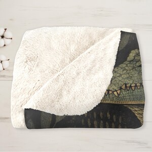 May include: A cozy, folded blanket with a cream-colored, plush sherpa lining and a dark patterned exterior. The exterior features a detailed illustration of a reptile's head in shades of green, brown, and black. The blanket is folded on a light wood surface.