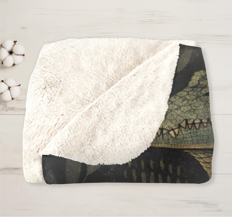 May include: A cozy, folded blanket with a cream-colored, plush sherpa lining and a dark patterned exterior. The exterior features a detailed illustration of a reptile's head in shades of green, brown, and black. The blanket is folded on a light wood surface.