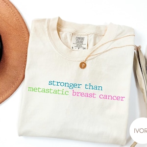 May include: An ivory Comfort Colors t-shirt with the text "stronger than metastatic breast cancer" in teal, green, and pink. A gold necklace with a pendant rests on the shirt. A brown hat is partially visible.