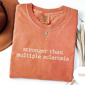 May include: A coral-colored t-shirt with the text "stronger than multiple sclerosis" in white. A gold necklace with a pendant rests on the shirt. A brown hat and dried plant are in the background.