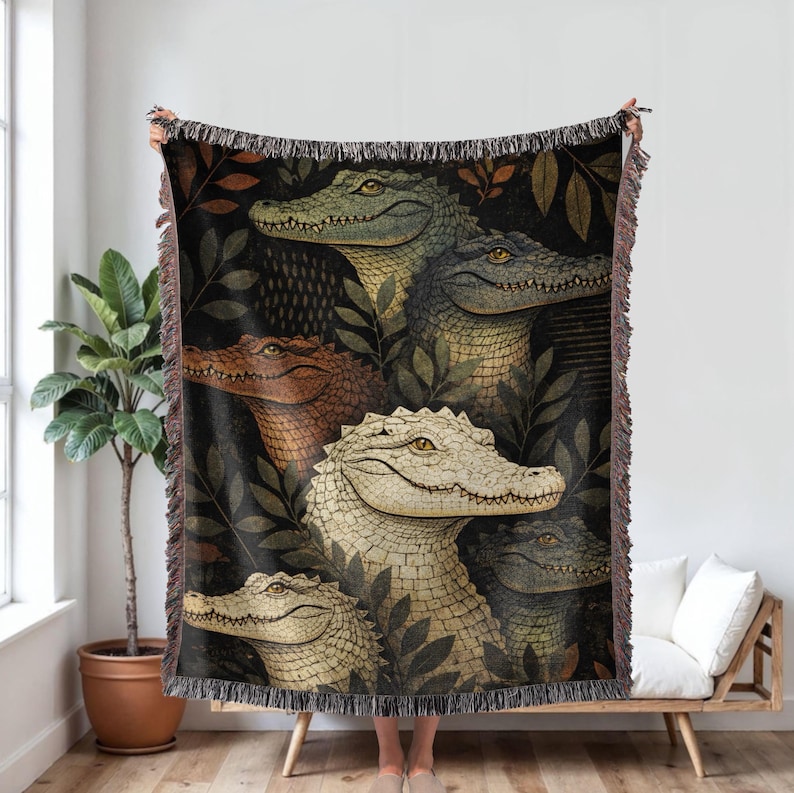 May include: A woven throw blanket featuring a detailed illustration of several alligators in various colors, including green, brown, and cream. The alligators are surrounded by dark green foliage against a black background. The blanket has a fringed edge.