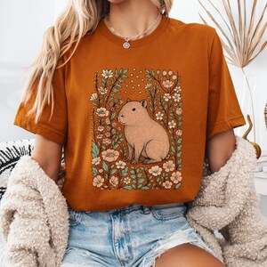 Puede incluir: Camiseta de color &oacute;xido con una ilustraci&oacute;n de carpincho rodeado de flores y follaje. El dise&ntilde;o incluye un carpincho marr&oacute;n claro, flores melocot&oacute;n y blancas y hojas verdes. La camiseta tiene mangas cortas y cuello redondo.