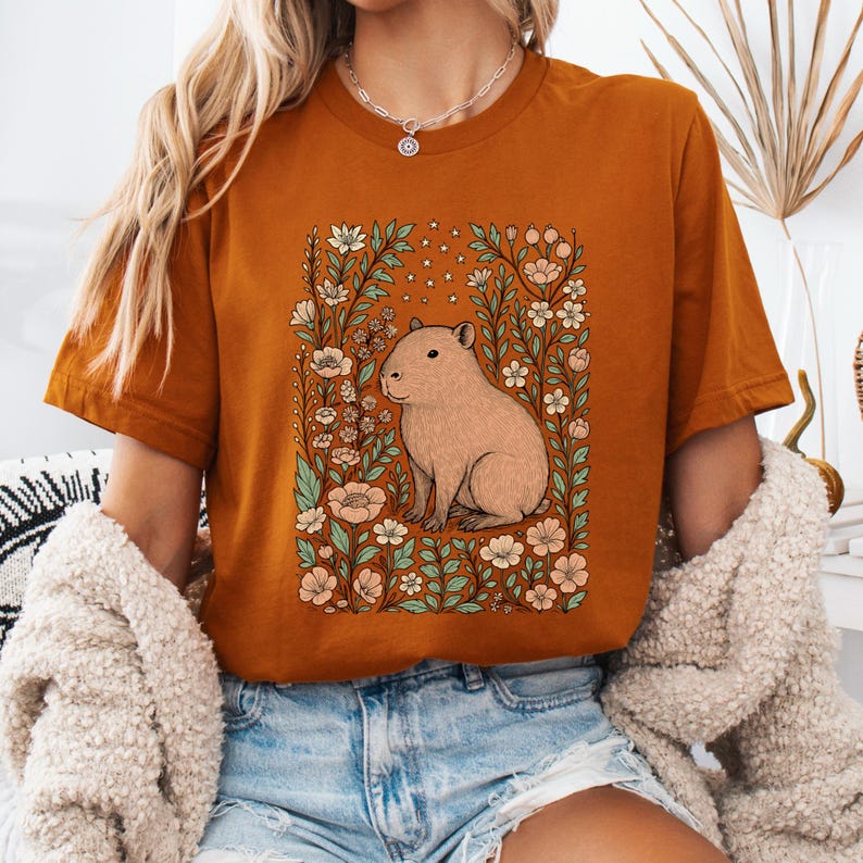 Puede incluir: Camiseta de color &oacute;xido con una ilustraci&oacute;n de carpincho rodeado de flores y follaje. El dise&ntilde;o incluye un carpincho marr&oacute;n claro, flores melocot&oacute;n y blancas y hojas verdes. La camiseta tiene mangas cortas y cuello redondo.