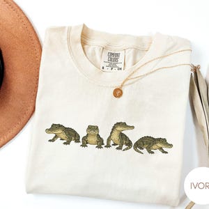 Op de afbeelding: Ivoor Comfort Colors T-shirt met vier gedetailleerde alligator illustraties in groen en bruin. Een gouden ketting met een hanger ligt op het shirt. Een bruine hoed en gedroogde planten zijn op de achtergrond.