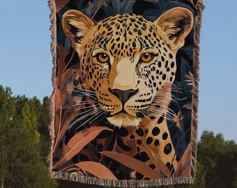 Op de afbeelding: Een geweven tapijtdeken met een close-up van een luipaardkop met een jungleachtergrond. De luipaard heeft bruine en zwarte vlekken en een lichtbruine neus. De achtergrond is donkerblauw met bruine en oranje bladeren.