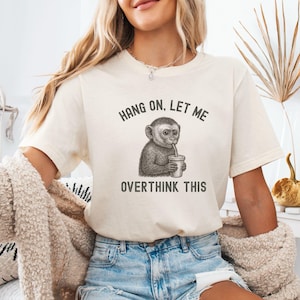 May include: Cream-colored t-shirt featuring a detailed illustration of a monkey sipping from a cup. The text "HANG ON, LET ME OVERTHINK THIS" is printed above and below the monkey graphic in a dark gray font. The shirt is made of a soft material.