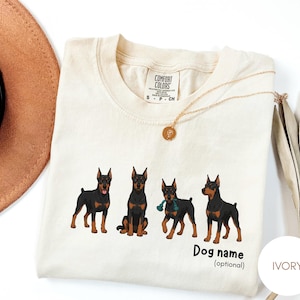 May include: An ivory-colored t-shirt featuring a graphic of four black and tan Doberman dogs in various poses. The text 'Dog name (optional)' is printed below the dogs. The shirt has a Comfort Colors brand label at the inner neckline.