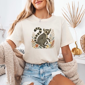 Könnte beinhalten: Cremefarbenes T-Shirt mit einer detaillierten Illustration eines Lemuren, umgeben von floralen Elementen. Der Lemur ist in Grautönen mit einem gestreiften Schwanz dargestellt. Das Design ist auf dem Shirt zentriert und schafft eine von der Natur inspirierte Ästhetik.