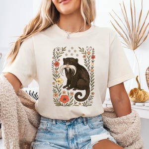 Könnte beinhalten: Cremefarbenes T-Shirt mit einer skurrilen Illustration eines dunkelbraunen Primaten mit langem weißem Schnurrbart, umrahmt von einem floralen Rand mit roten, gelben und weißen Blumen. Das Shirt hat kurze Ärmel und eine lockere Passform.