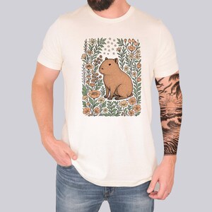 Puede incluir: Camiseta color crema con una ilustraci&oacute;n de carpincho marr&oacute;n rodeada de flores en tonos naranja, verde y crema. El carpincho est&aacute; centrado dentro de un marco floral. La camiseta la lleva una persona con vaqueros azules.