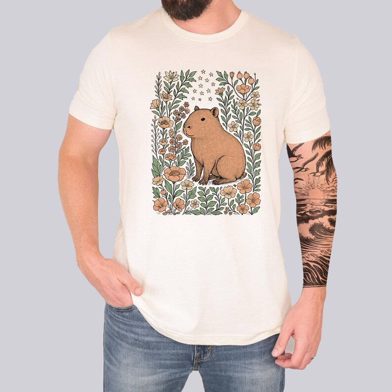 Puede incluir: Camiseta color crema con una ilustraci&oacute;n de carpincho marr&oacute;n rodeada de flores en tonos naranja, verde y crema. El carpincho est&aacute; centrado dentro de un marco floral. La camiseta la lleva una persona con vaqueros azules.
