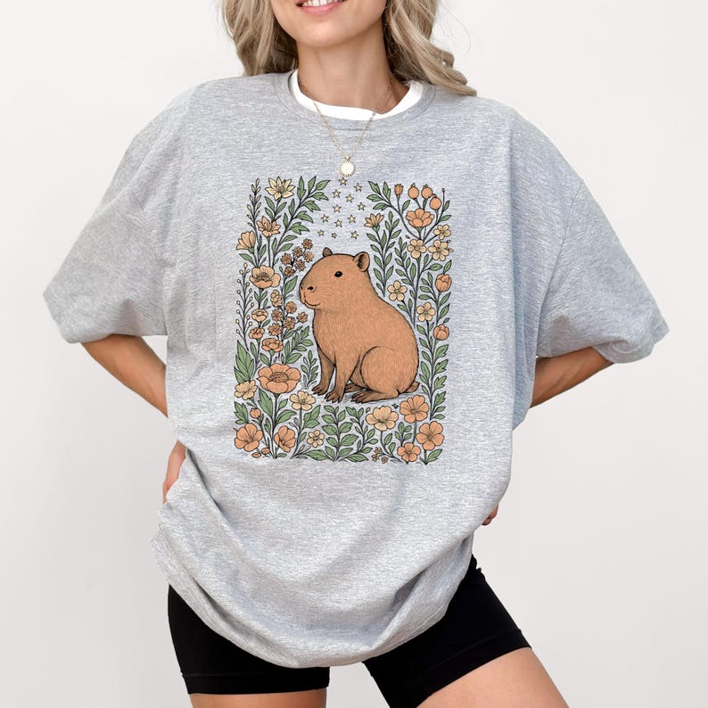 Puede incluir: Una sudadera gris claro con una ilustraci&oacute;n de carpincho rodeada de flores y estrellas. El carpincho es marr&oacute;n, y las flores son melocot&oacute;n y amarillas. La sudadera tiene cuello redondo y mangas largas.