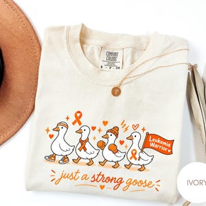 May include: An ivory t-shirt featuring a graphic of four white geese in orange accessories. The geese wear headbands, scarves, and boxing gloves, with one holding an orange flag that reads Leukemia Warrior. Text below reads just a strong goose.