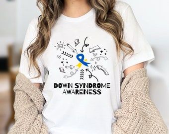 Puede incluir: Camiseta blanca con un diseño gráfico que promueve la concienciación sobre el síndrome de Down. El diseño incluye una cinta azul y amarilla, flechas y las palabras "DOWN SYNDROME AWARENESS" en negro. La camiseta es de material suave.