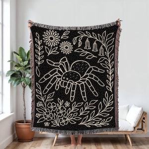 Puede incluir: Manta tejida negra con un diseño de araña y flores crema. La manta tiene un borde con flecos y se exhibe en interiores. El diseño incluye flores, hojas y una araña grande.