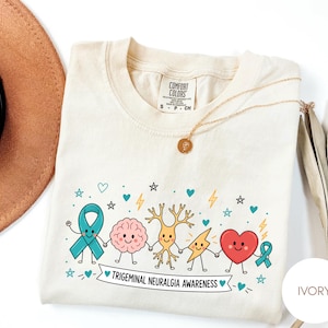 May include: An ivory t-shirt featuring a graphic of a teal awareness ribbon, a brain, a neuron, a lightning bolt, and a heart holding hands. Below the characters, a banner displays the text "TRIGEMINAL NEURALGIA AWARENESS" with small stars and hearts.