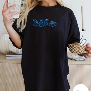 May include: Black t-shirt featuring a row of five blue frog illustrations. The frogs are detailed with shading and positioned horizontally across the chest. The shirt is a solid black color and appears to be made of a soft material.