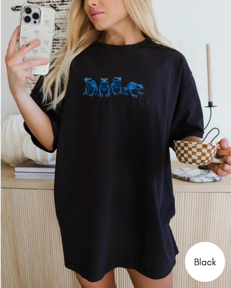 May include: Black t-shirt featuring a row of five blue frog illustrations. The frogs are detailed with shading and positioned horizontally across the chest. The shirt is a solid black color and appears to be made of a soft material.