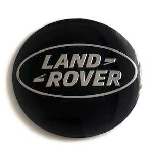Puede incluir: Un emblema negro y circular con el logotipo de Land Rover en plata. El logotipo, en forma ovalada, presenta las palabras "LAND" y "ROVER" en una fuente sans-serif. El emblema tiene un acabado brillante.