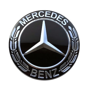 Peut inclure: Un logo Mercedes-Benz argenté et noir avec les mots "Mercedes" et "Benz" dans un cercle argenté autour d'une étoile à trois branches.