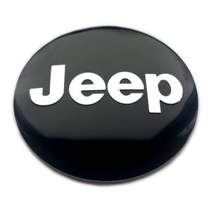 Może przedstawiać: Błyszcząca, czarna, okrągła odznaka z białym napisem "Jeep". Napis jest pogrubiony i bezszeryfowy. Odznaka ma lekko wypukły kształt i odblaskową powierzchnię, tworząc kontrast między czarnym tłem a białym tekstem.