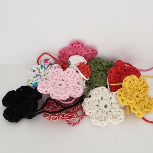 May include: A collection of crocheted flower shapes in various colors, including pink, red, yellow, green, white, and black. The flowers are arranged in a pile on a white surface.