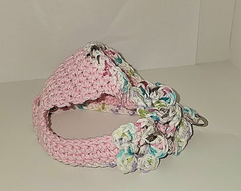 May include: A handmade crochet headband in light pink with a white and multicolored floral accent. The headband is crafted with a textured stitch and features a circular design. The floral accent includes shades of pink, blue, and green.