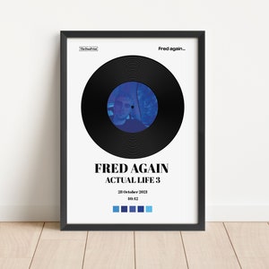 May include: A framed poster with a black border features a black vinyl record design with a blue center image of a person. The poster reads "FRED AGAIN ACTUAL LIFE 3" with the date "28 October 2021" and time "10:12".