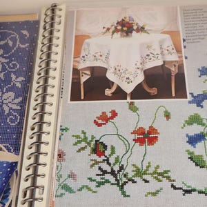 Op de afbeelding: Pagina uit een knutselboek met kruissteekpatronen. Het toont een foto van een wit tafelkleed met bloemenborduurwerk en een kruissteekontwerp van rode klaprozen en blauwe bloemen. Aan de linkerkant is een blauw-wit bloemenpatroon te zien.