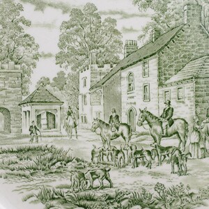 May include: A decorative plate with a green and white transferware design. The scene depicts a rural landscape with buildings, trees, people on horseback, and dogs. The plate has a floral border.