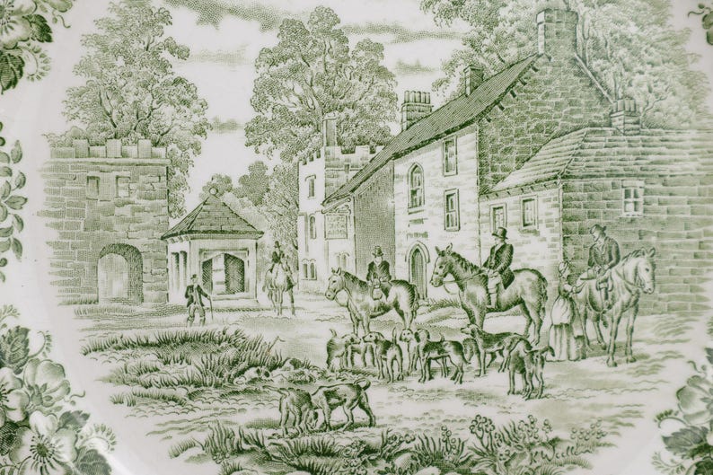 May include: A decorative plate with a green and white transferware design. The scene depicts a rural landscape with buildings, trees, people on horseback, and dogs. The plate has a floral border.