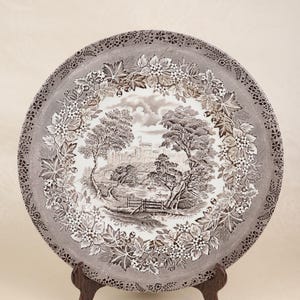 May include: A decorative plate with a detailed landscape scene in shades of brown and white. The plate features a central image of a castle, trees, and a river, surrounded by a floral border. The plate is displayed on a wooden stand.