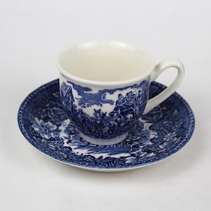 Puede incluir: Juego de taza y platillo de té de cerámica azul y blanco. La taza y el platillo presentan un diseño de transferencia detallado que representa una escena rural y motivos florales. El juego se muestra sobre un fondo blanco.