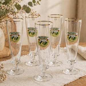 Könnte beinhalten: Fünf klare Ayinger Biergläser mit goldenen Rändern und einem Logo. Die Gläser stehen auf einem weißen Tuch auf einem Holztisch. Die Gläser haben ein Fassungsvermögen von 16,0. Eine Vase mit Blumen befindet sich im Hintergrund.