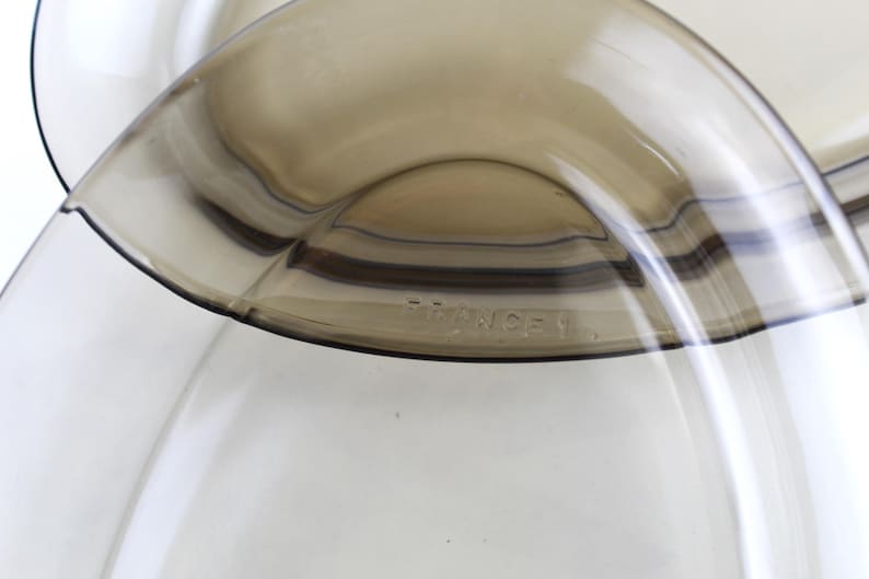 May include: Close-up of two overlapping, transparent, brown-tinted glass bowls. The bowls have a curved, organic shape. The text "FRANCE 1" is visible on the rim of one bowl. The bowls are set against a white background.
