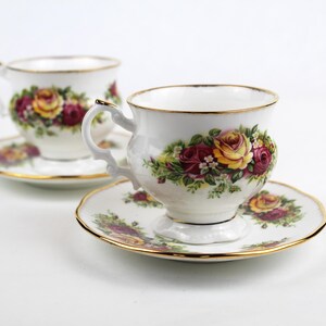 May include: Two white teacups and saucers with gold trim. Each cup and saucer features a floral design with red and yellow roses and green leaves. The teacups have ornate handles.