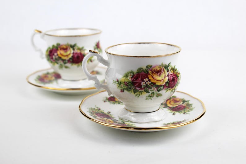 May include: Two white teacups and saucers with gold trim. Each cup and saucer features a floral design with red and yellow roses and green leaves. The teacups have ornate handles.