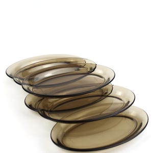May include: A stack of five oval-shaped, transparent brown glass plates. The plates have a slightly raised rim and a smooth surface. The plates are stacked at a slight angle, showcasing their shape and color against a white background.