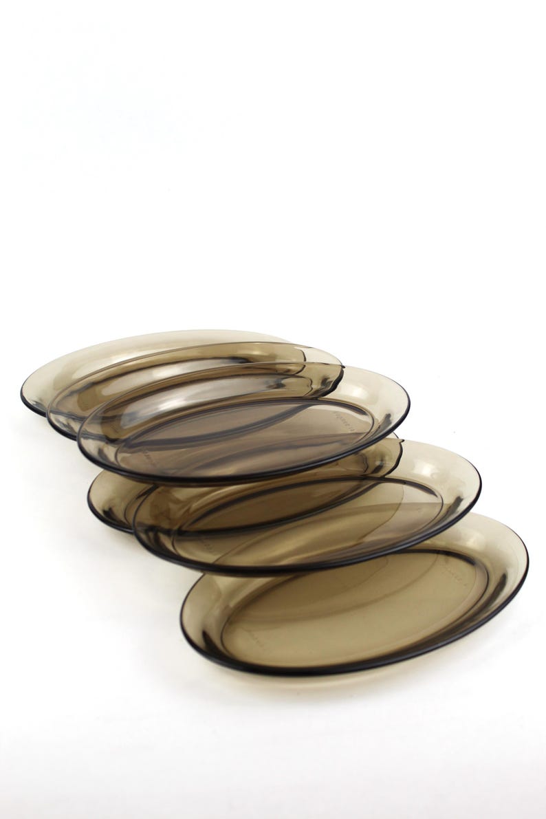 May include: A stack of five oval-shaped, transparent brown glass plates. The plates have a slightly raised rim and a smooth surface. The plates are stacked at a slight angle, showcasing their shape and color against a white background.