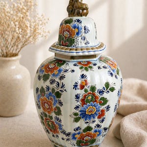Könnte beinhalten: Ein weißer Ingwertopf aus Keramik mit abnehmbarem Deckel und einem braunen, skulptierten Knauf. Der Topf ist mit einem sich wiederholenden Muster aus blauen, orangefarbenen und grünen Blumenmotiven verziert. Der Körper hat eine vertikale Rippenstruktur.