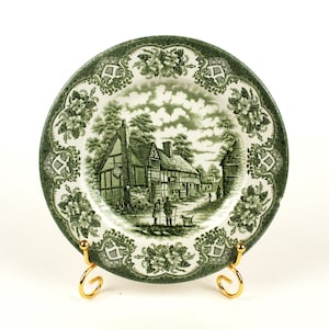 Peut inclure: Assiette décorative verte et blanche représentant un paysage rural. L'assiette a une bordure détaillée avec des motifs floraux et géométriques. L'assiette est présentée sur un support en métal doré.