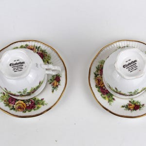 May include: Two white teacups and saucers with a floral rose pattern in shades of red, yellow, and green. The rims are trimmed with gold. The teacups are upside down, revealing the "Elizabethan" stamp on the bottom.