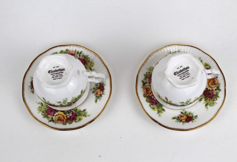May include: Two white teacups and saucers with a floral rose pattern in shades of red, yellow, and green. The rims are trimmed with gold. The teacups are upside down, revealing the "Elizabethan" stamp on the bottom.