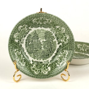 Może przedstawiać: Zielono-biały talerz dekoracyjny z motywem krajobrazu, prezentowany na złotym stojaku. W tle widoczna pasująca miska. Talerz przedstawia szczegółową ilustrację budynku, drzew i wody.