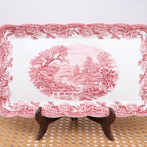 May include: A rectangular serving platter with a white base and a red floral and landscape design. The platter has a scalloped edge and is supported by a dark brown wooden stand. The central design depicts a landscape scene.