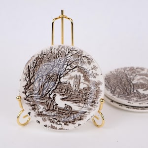 May include: A set of three small round ceramic plates featuring a brown and white transferware landscape scene with a river, trees, and a building. One plate is displayed upright on a gold-toned metal wire stand, while two others are stacked.