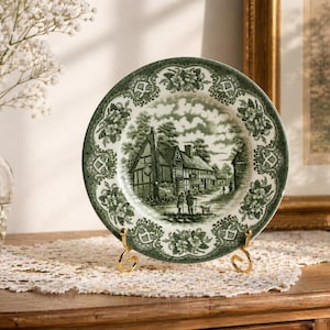 May include: A decorative plate with a green and white landscape design. The plate is displayed on a gold-coloured stand. The plate features a detailed illustration of a building and people. The plate is on a wooden surface.