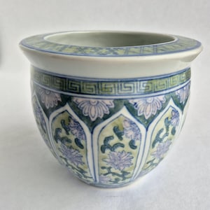 Puede incluir: Un macetero de cerámica blanca con un diseño floral azul y verde. El macetero tiene un borde ancho y un cuerpo redondeado.