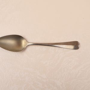 Puede incluir: Una cuchara plateada con un cuenco redondeado y un mango largo y delgado. La cuchara está hecha de un metal brillante y está colocada sobre un fondo de color claro. La cuchara es un utensilio común para comer.