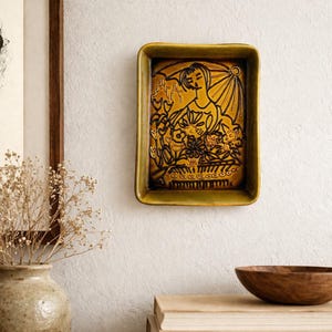 May include: A rectangular ceramic wall plaque in a deep mustard yellow glaze. The surface features a relief design of a person holding an umbrella over a collection of flowers, rendered in dark brown outlines against the glossy yellow background.